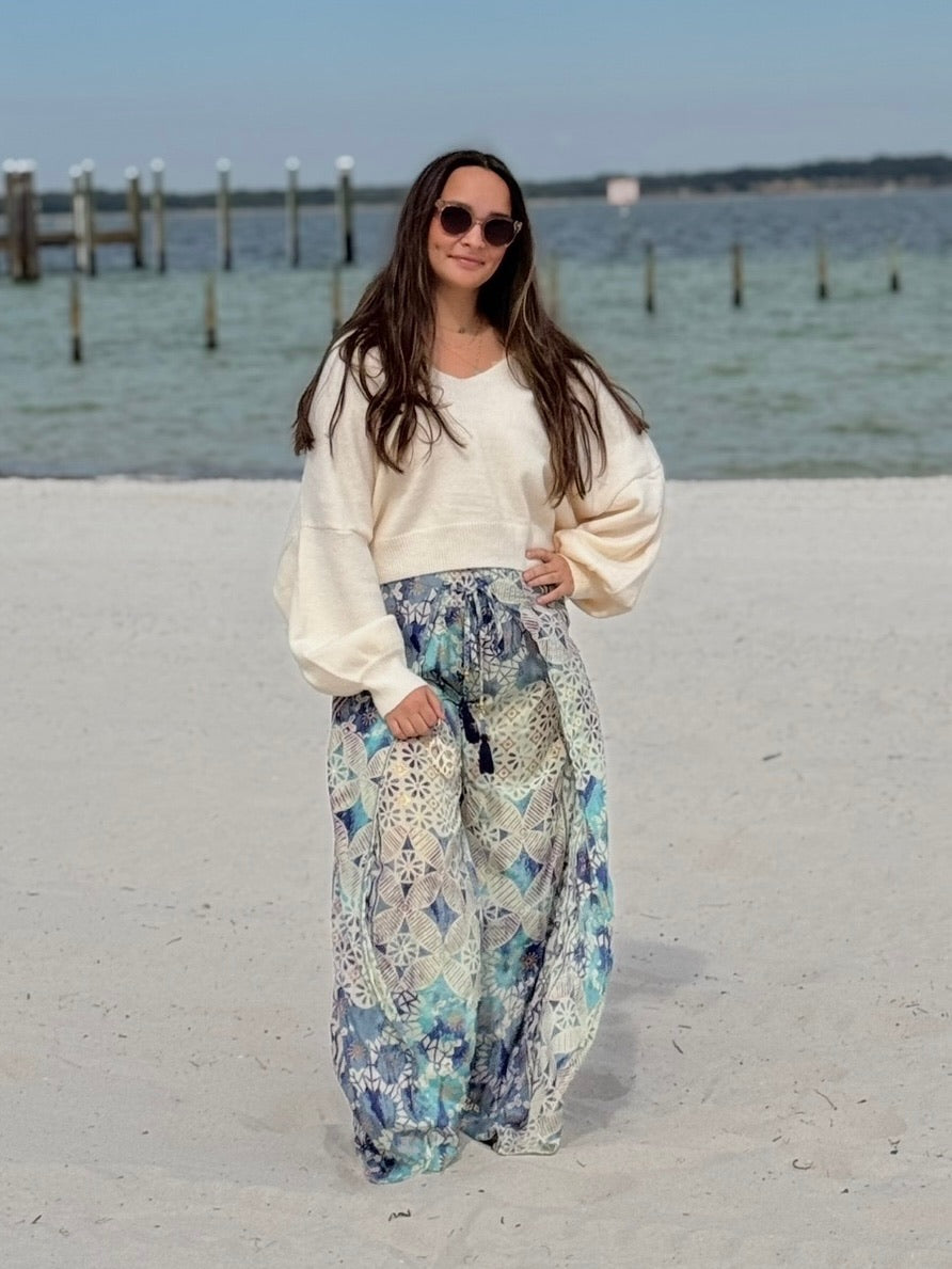 Woman in a white sweater and floral flowy pants standing on a sandy beach with water and dock in the background.