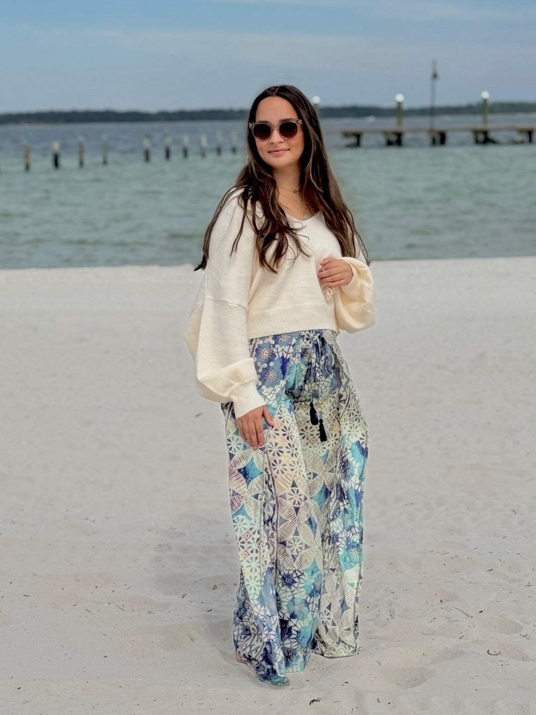 Woman in a white sweater and floral pants standing on a sandy beach with water and a dock in the background.