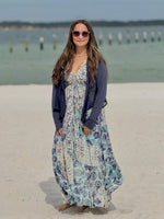 Woman in a floral dress and navy cardigan standing on a sandy beach with water and wooden posts in the background.