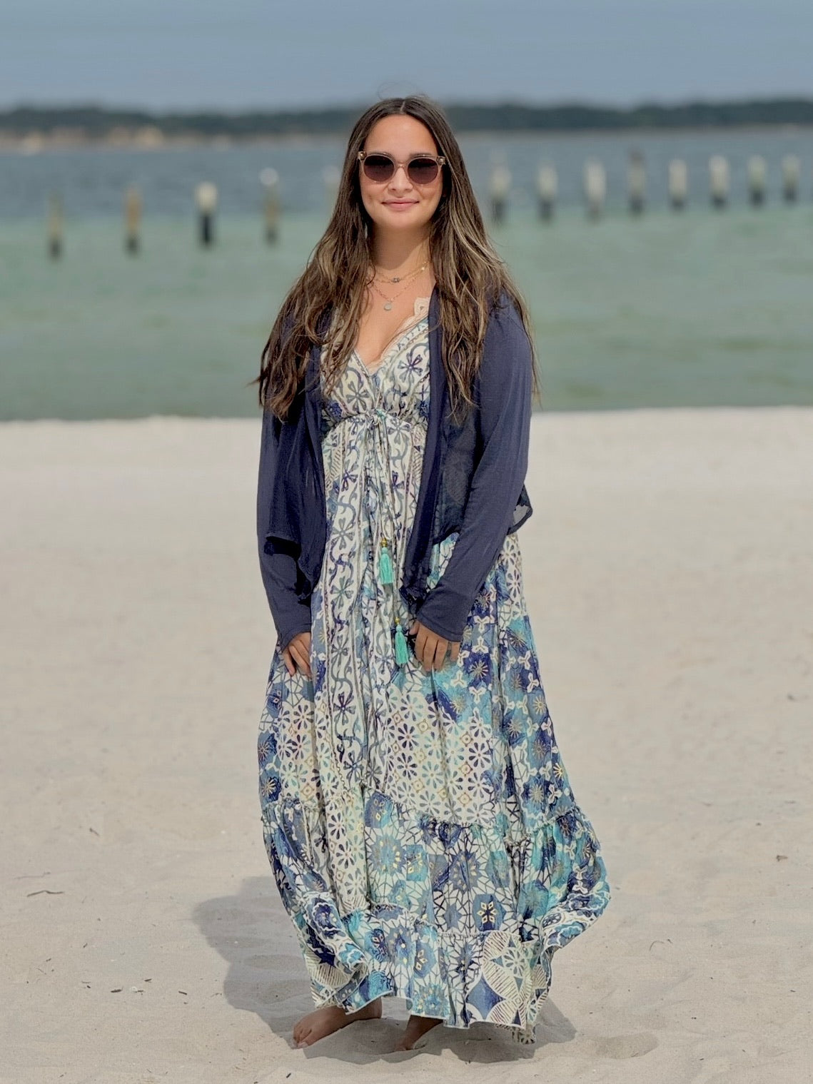 Woman in a floral dress and navy cardigan standing on a sandy beach with water and wooden posts in the background.