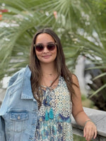 Woman wearing sunglasses and a denim jacket with a floral dress, standing outdoors.