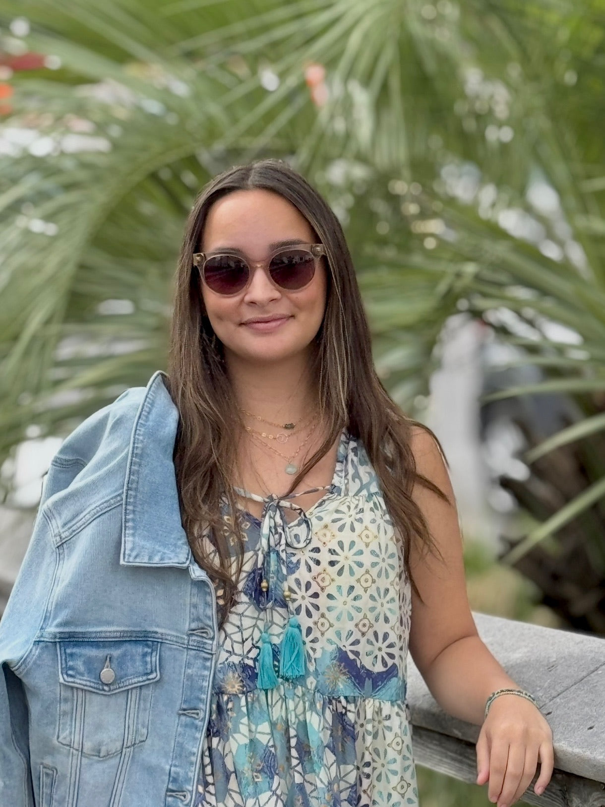 Woman wearing sunglasses and a denim jacket with a floral dress, standing outdoors.