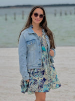 Woman in a floral dress and denim jacket standing on a beach.