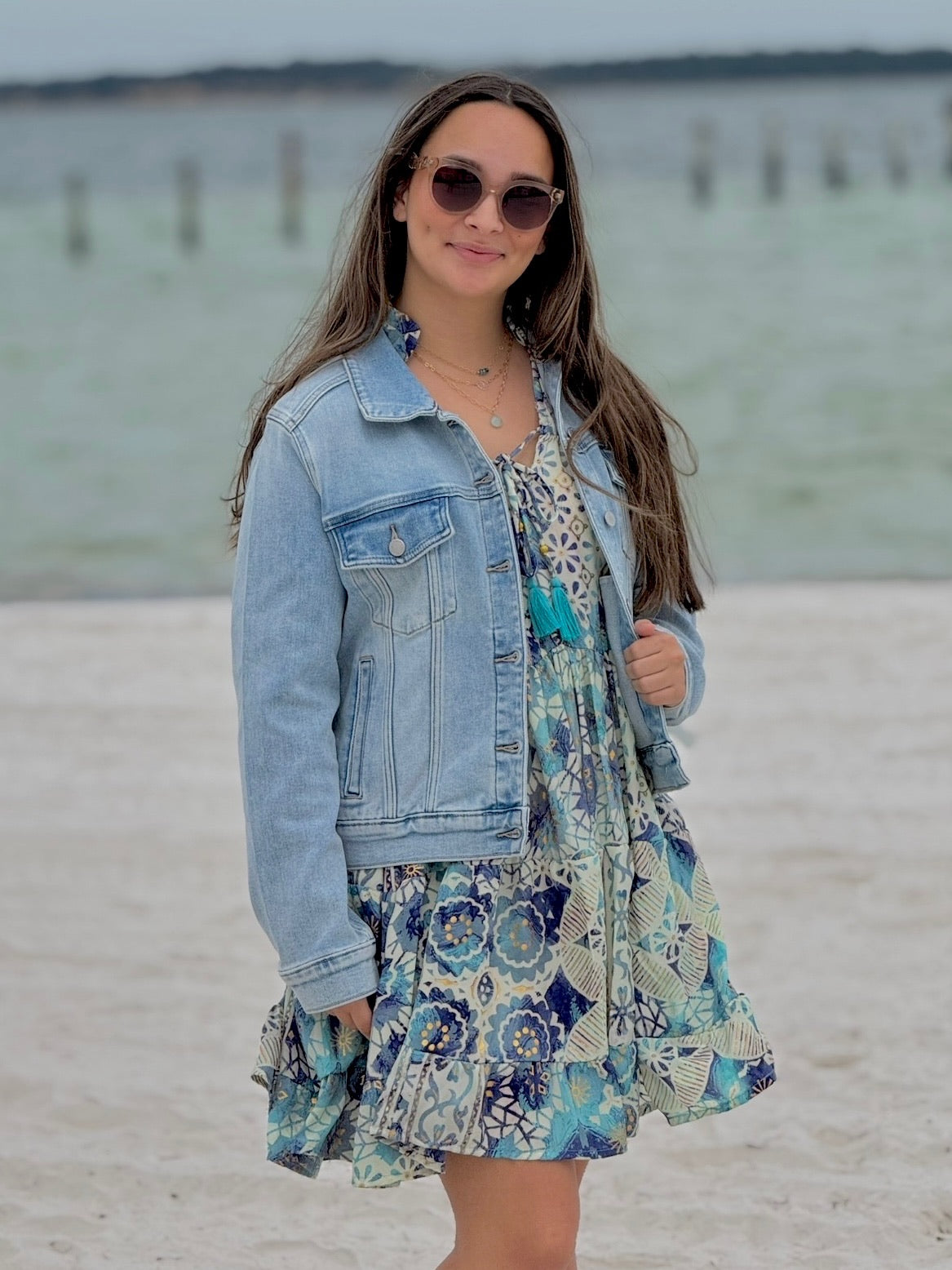 Woman in a floral dress and denim jacket standing on a beach.