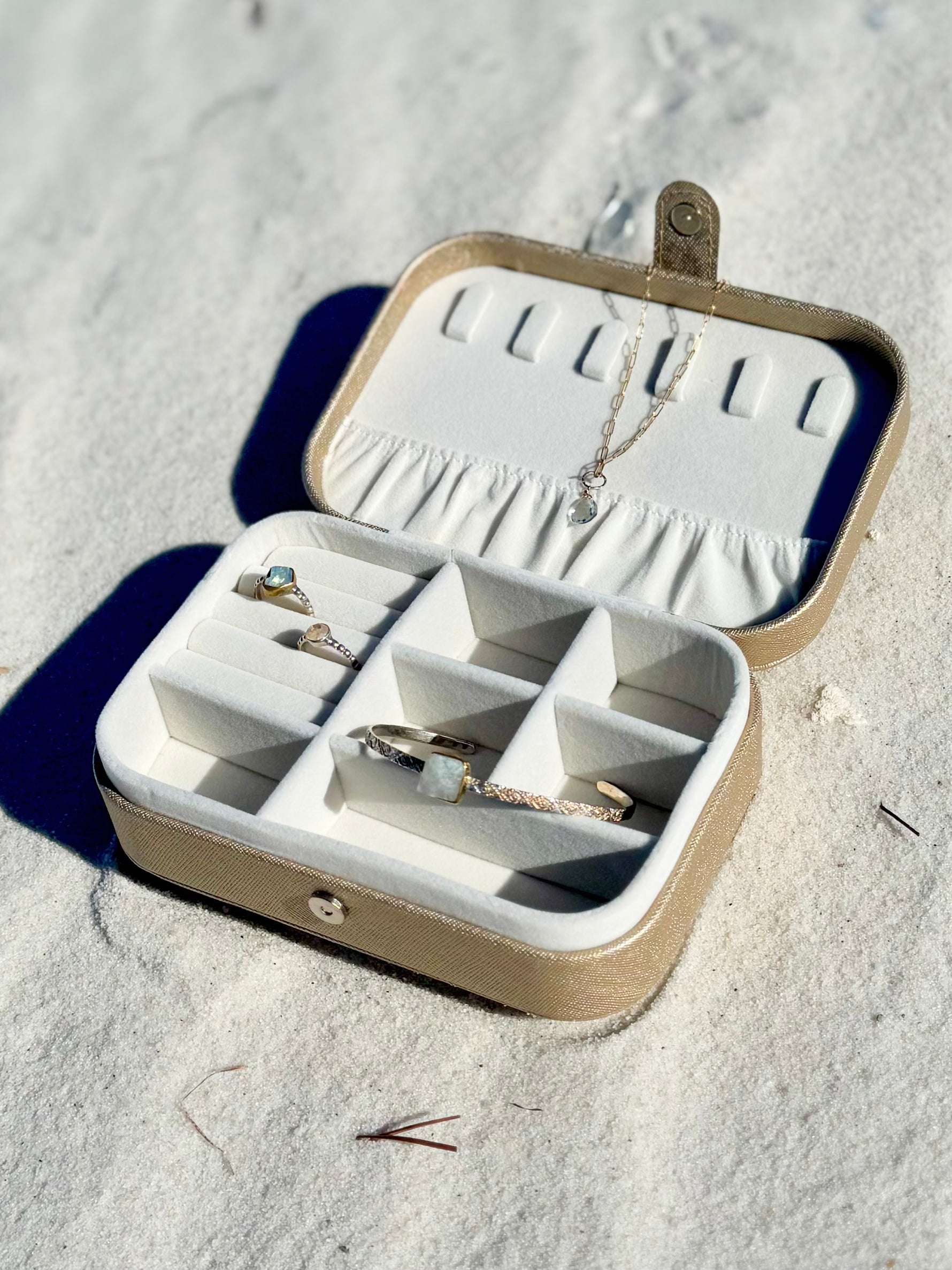 Elizabeth Jewelry Case in gold resting on white sand, displaying jewelry arranged inside the soft velvet compartments.