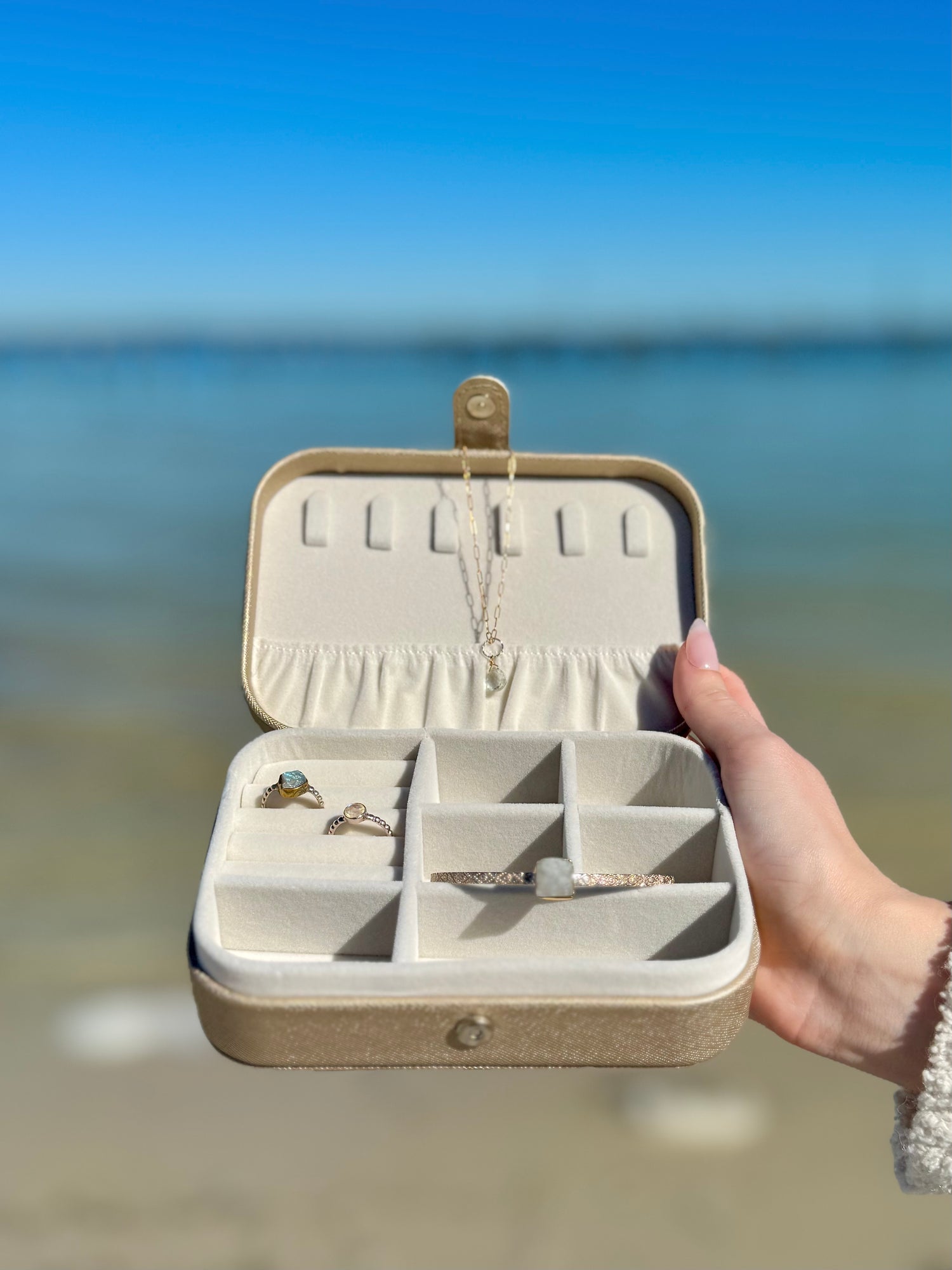 Open Elizabeth Jewelry Case in gold held near the ocean displaying rings, a bracelet, and a necklace inside the soft velvet compartments.
