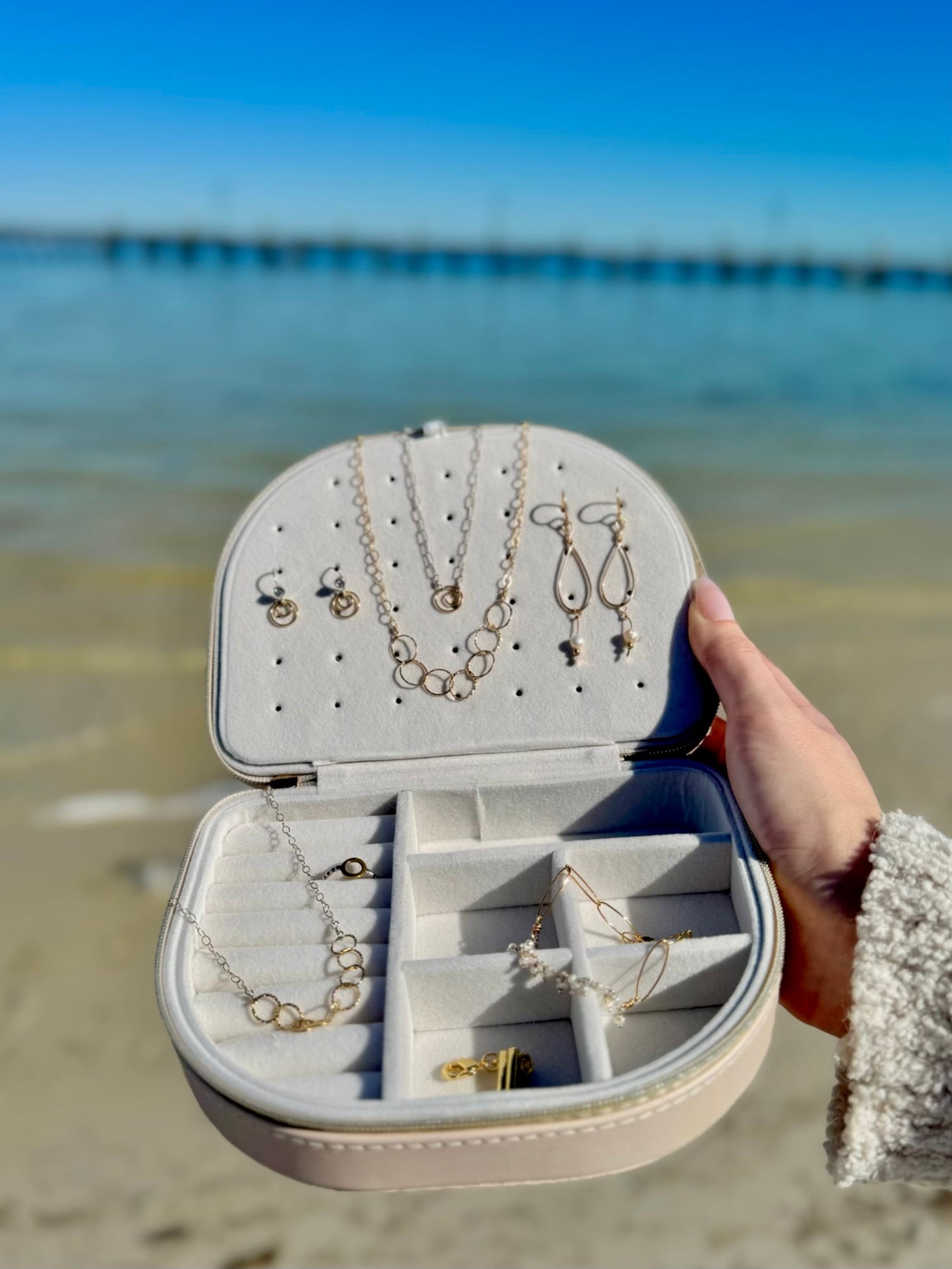 Open Victoria Curved Jewelry Case held by the ocean, displaying organized necklaces, earrings, and bracelets inside the soft velvet compartments.