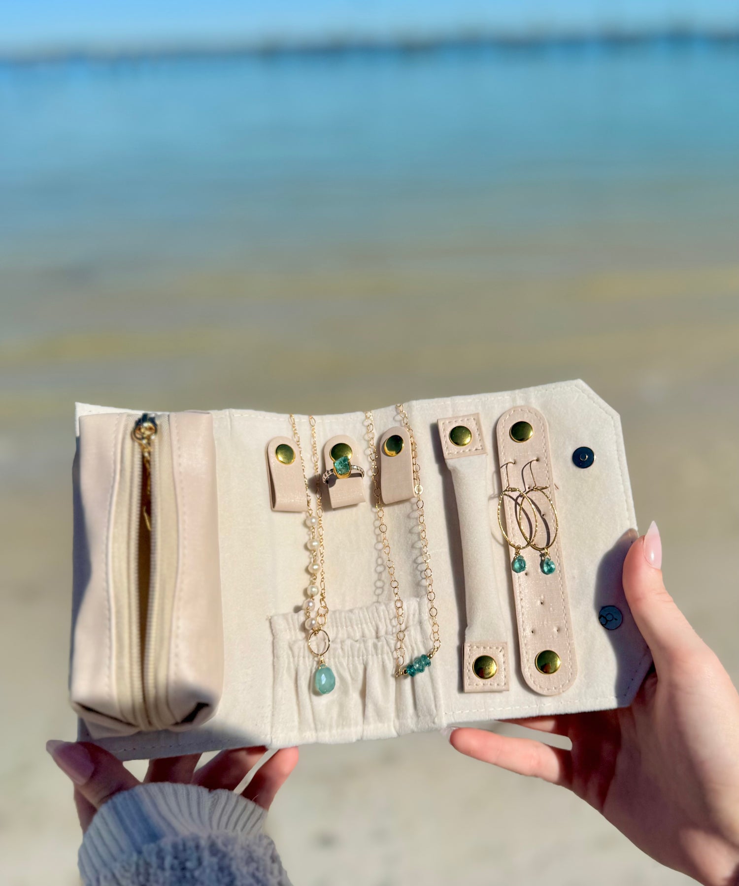 Pink Nina Halls jewelry roll-up case held open by the ocean, displaying organized necklaces and earrings inside.