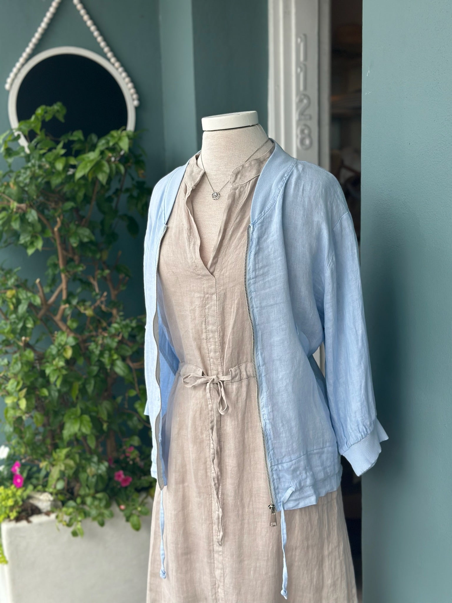 Light blue linen zip-front jacket with ribbed collar and cuffs, styled over a neutral linen dress. Features a relaxed fit, lightweight fabric, side pockets, and adjustable drawstring hem details.
