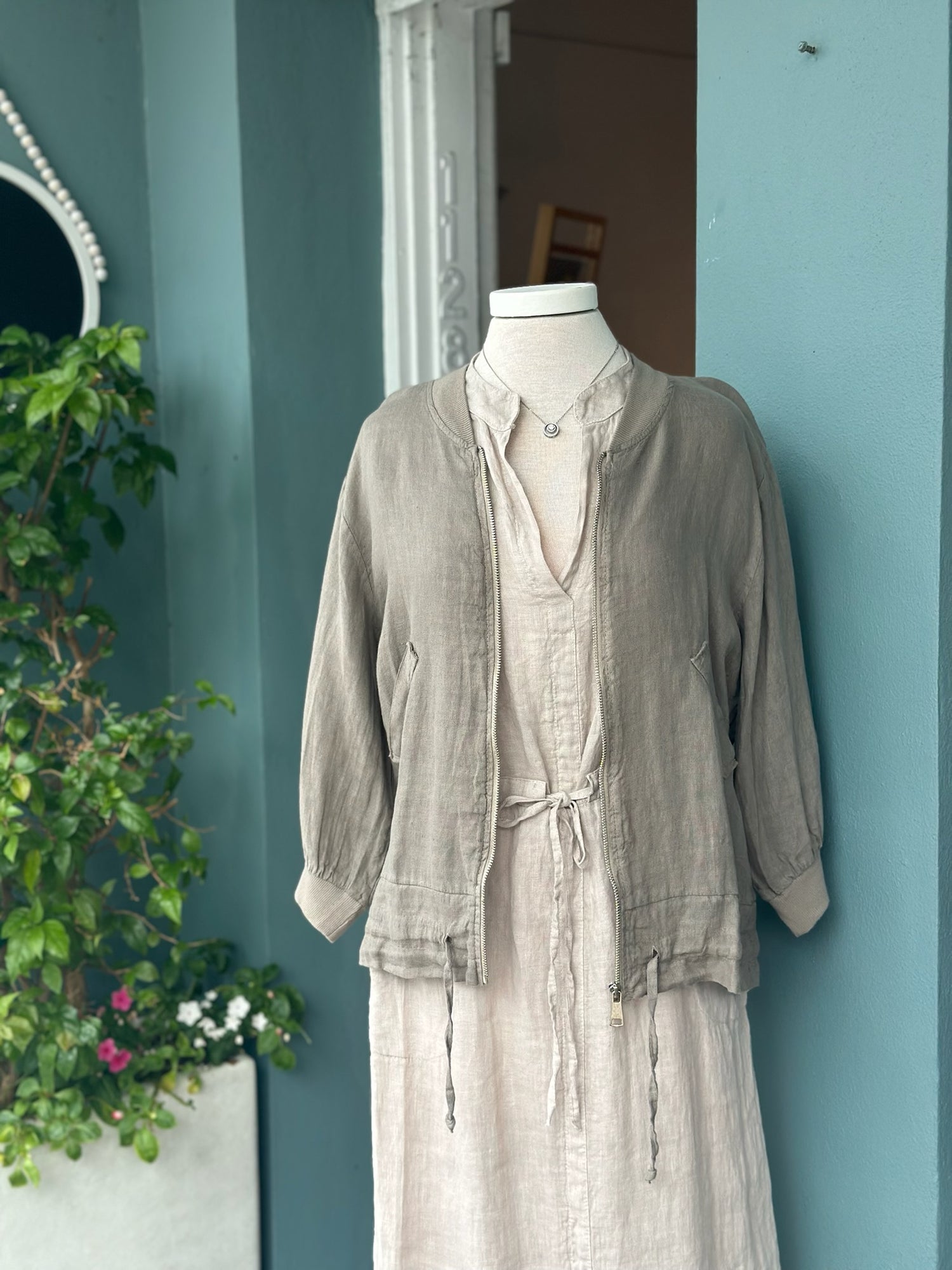 Taupe linen zip-front jacket with ribbed collar and cuffs, side pockets, and adjustable drawstring hem details, layered over a natural linen dress.