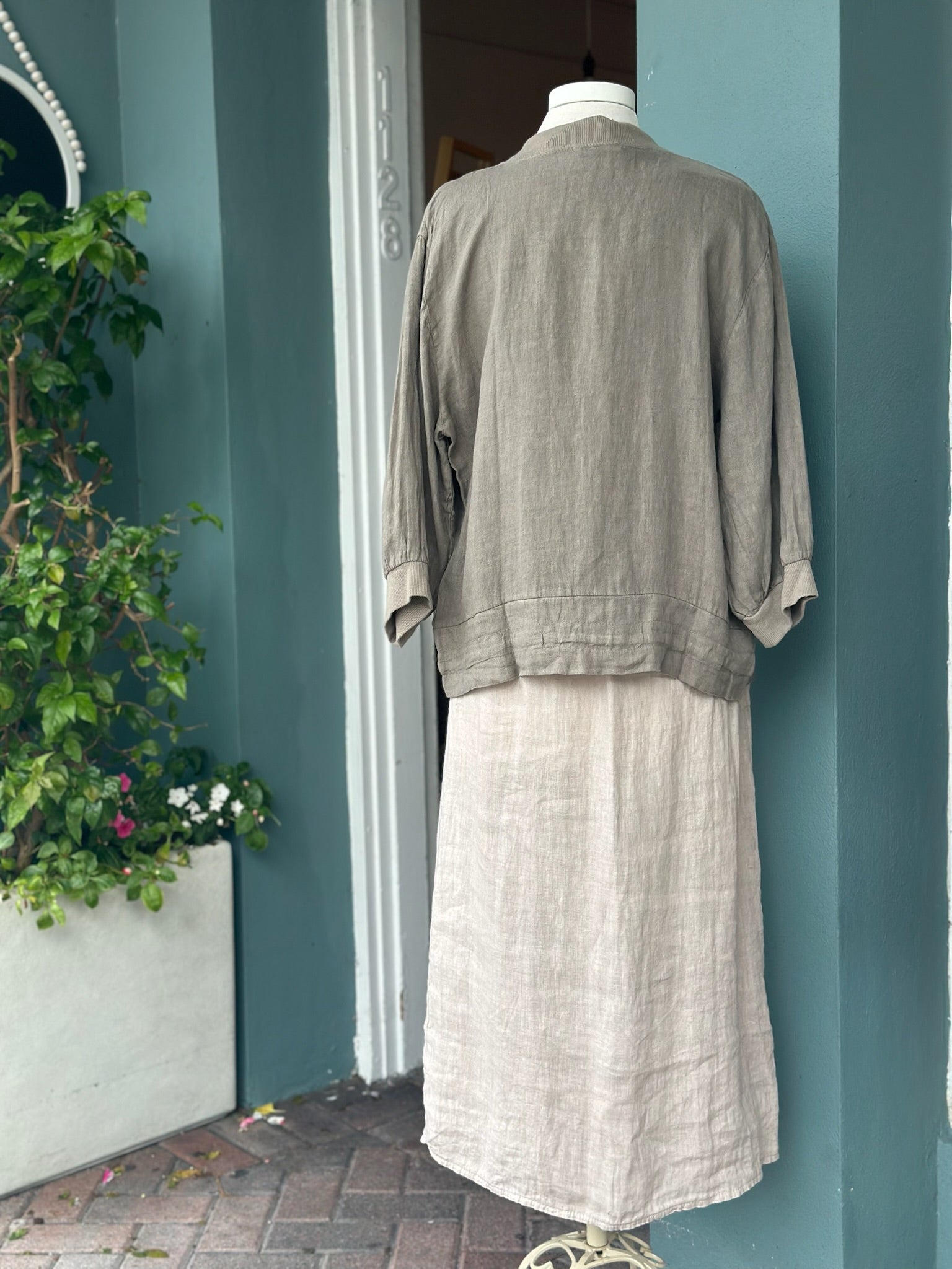 Back view of a taupe linen jacket featuring a relaxed silhouette, ribbed collar and cuffs, and lightweight linen fabric, styled over a natural linen dress.
