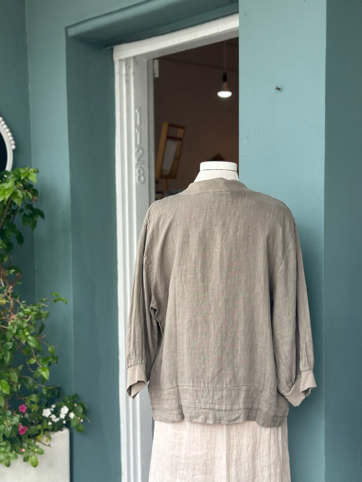Back view of a taupe linen jacket featuring a relaxed silhouette, ribbed collar and cuffs, and lightweight linen fabric, styled over a natural linen dress.