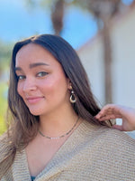 Woman with long brown hair wearing earrings and a necklace, outdoors.