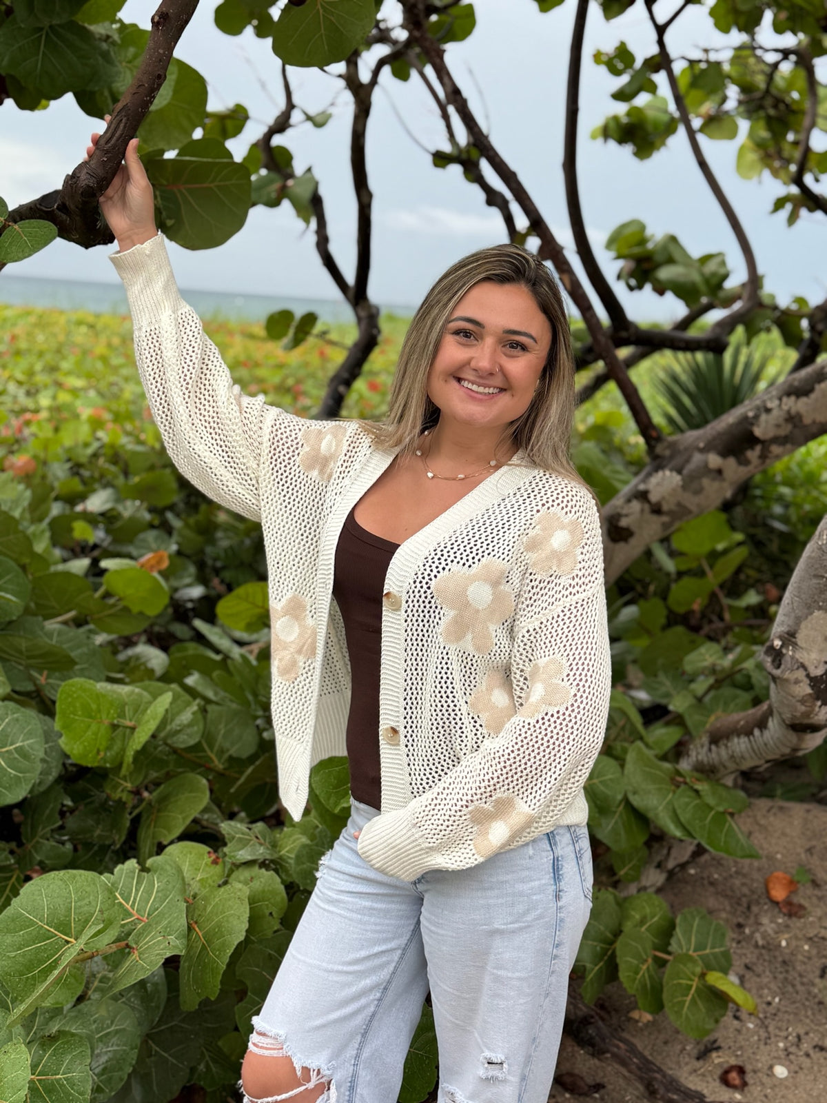 Woman wearing an ivory open-knit floral cardigan with beige flower details, brown tank top, and light wash distressed jeans near the beach, Ardor Boutique.