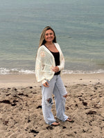 Woman modeling an ivory open-knit button-front floral cardigan with beige flower details near the ocean, Ardor Boutique.
