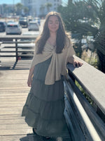 Woman in a layered dress with a beige shawl standing on a wooden deck with a cityscape background.