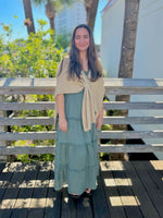 Woman in a green dress with a beige shawl standing on a wooden deck with trees and a building in the background.