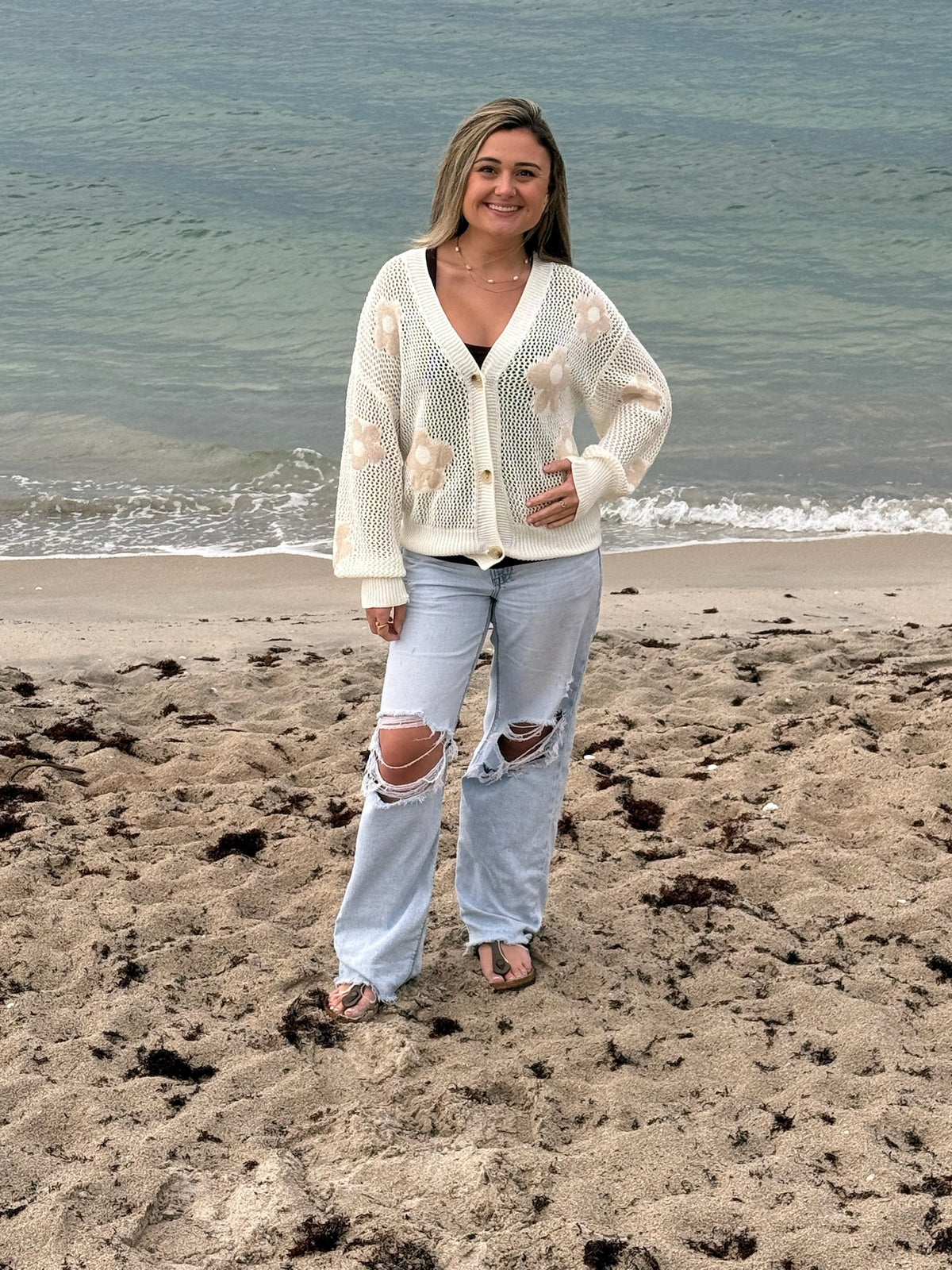 Woman wearing an ivory floral open-knit cardigan layered over a brown tank with light wash distressed wide-leg jeans on the beach, Ardor Boutique.