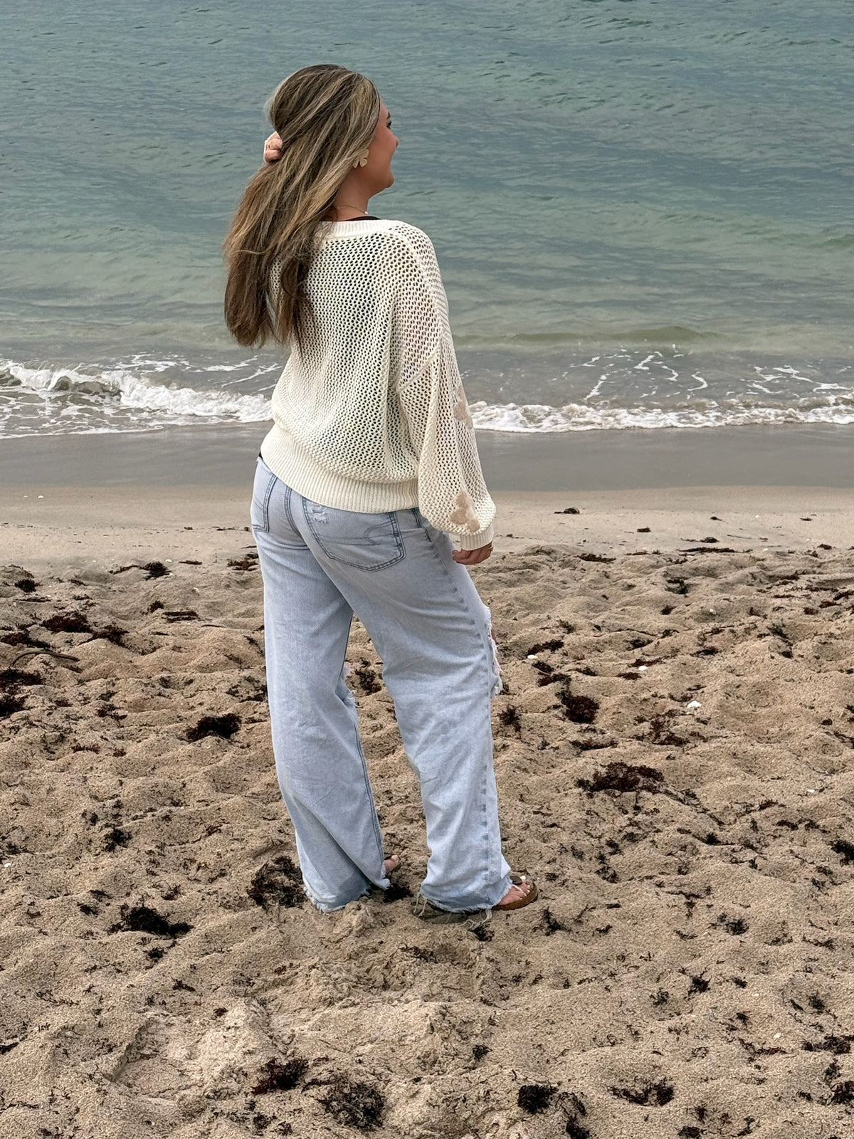 Back view of an ivory open-knit floral cardigan with ribbed trim, styled with light wash wide-leg jeans on the beach, Ardor Boutique.