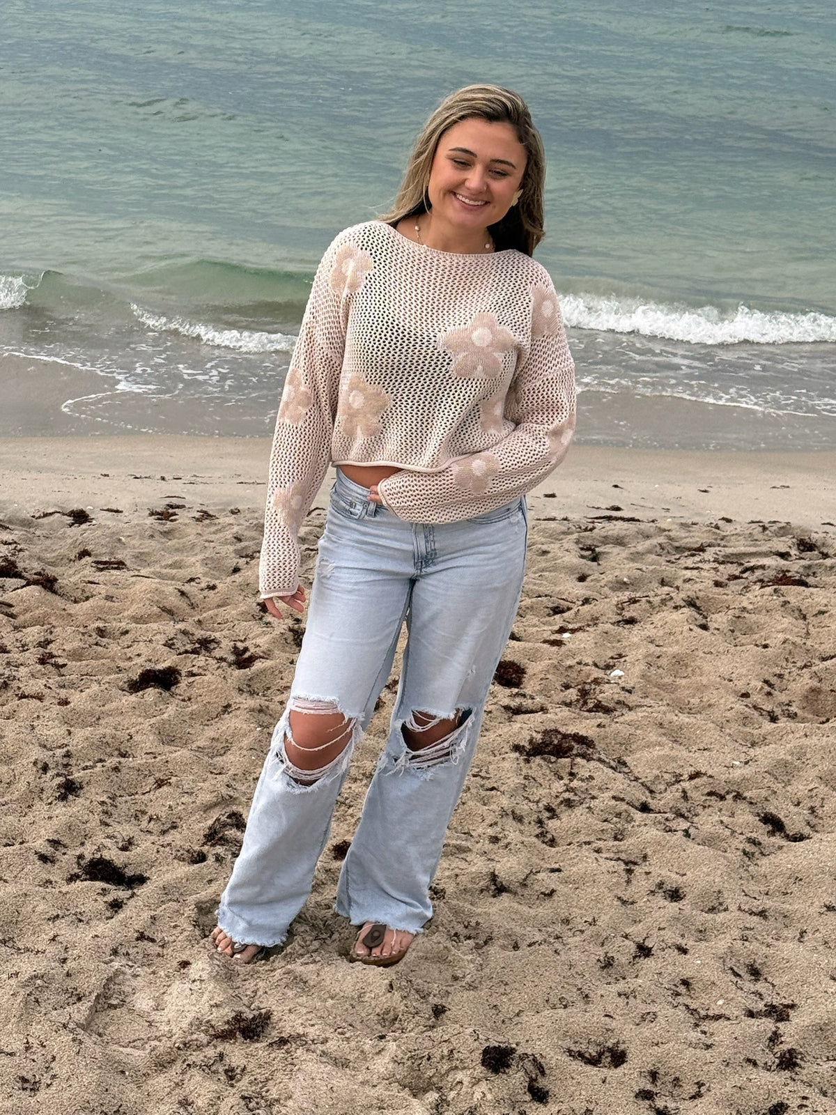 Woman wearing a beige open-knit floral sweater with light wash distressed wide-leg jeans on the beach by the ocean, Ardor Boutique.
