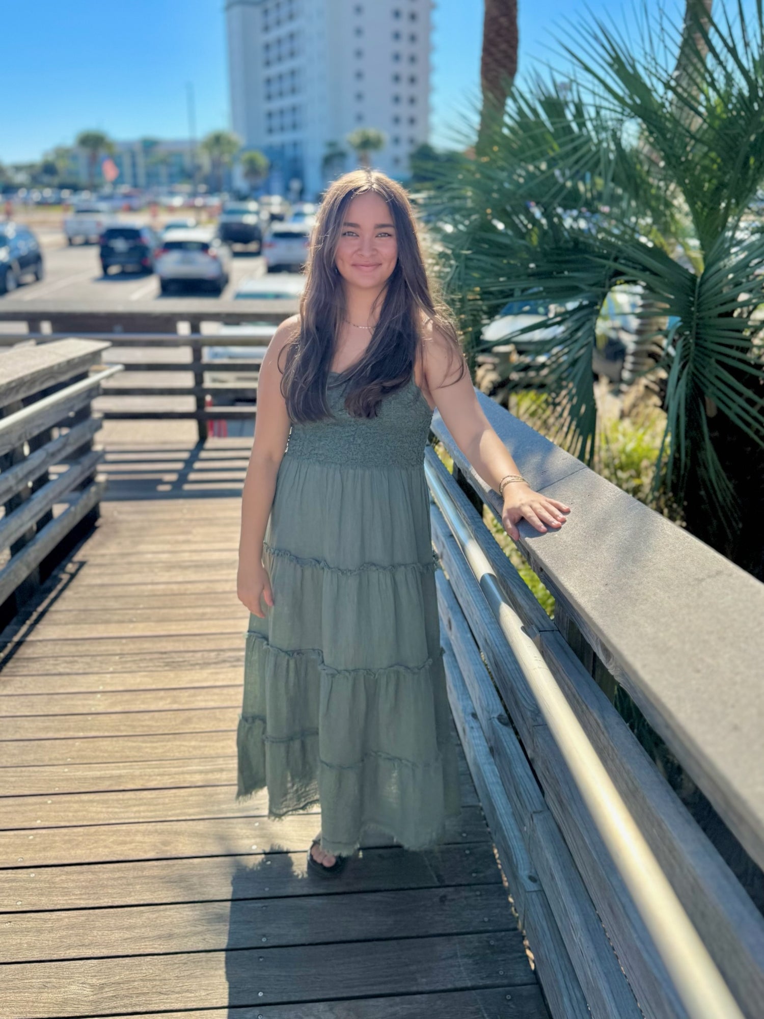 Woman in a green dress standing on a wooden boardwalk with palm trees and vehicles in the background.