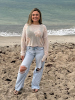 Woman wearing a beige open-knit floral sweater with light wash distressed wide-leg jeans by the ocean, Ardor Boutique.