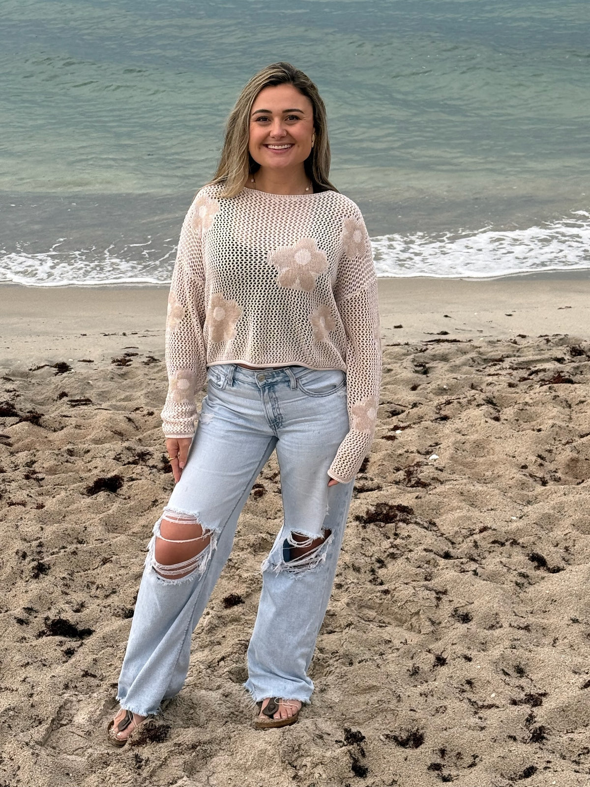 Woman wearing a beige open-knit floral sweater with light wash distressed wide-leg jeans by the ocean, Ardor Boutique.