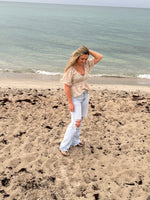 Woman wearing a taupe and ivory floral smocked blouse with flutter sleeves and light wash distressed jeans on the beach, Ardor Boutique.