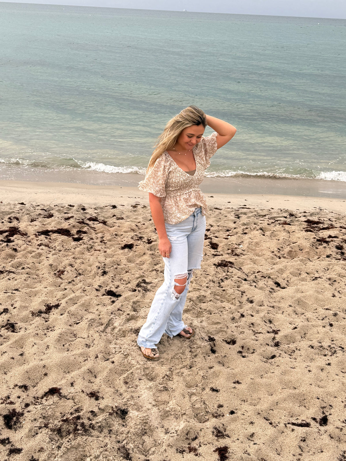 Woman wearing a taupe and ivory floral smocked blouse with flutter sleeves and light wash distressed jeans on the beach, Ardor Boutique.