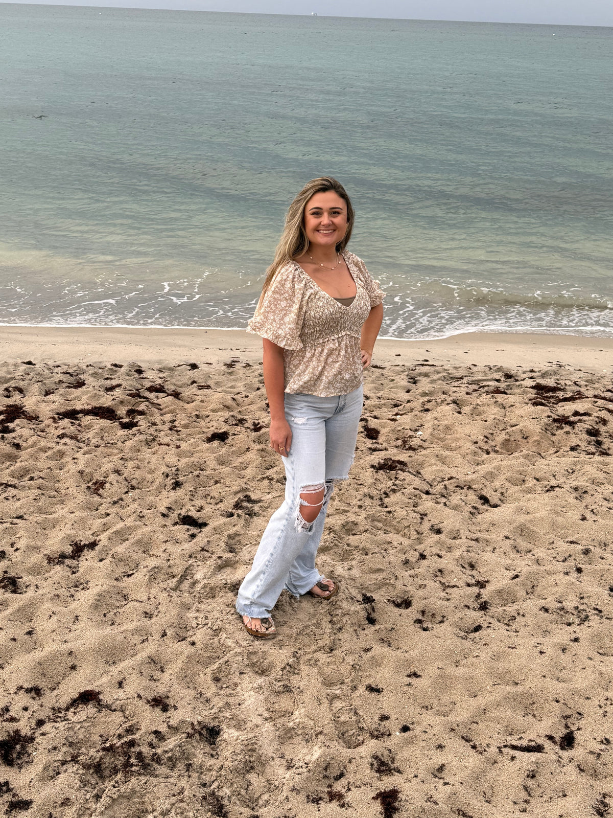 Woman modeling a taupe floral smocked blouse with a sweetheart neckline, flutter sleeves, and peplum-style hem by the ocean, Ardor Boutique.