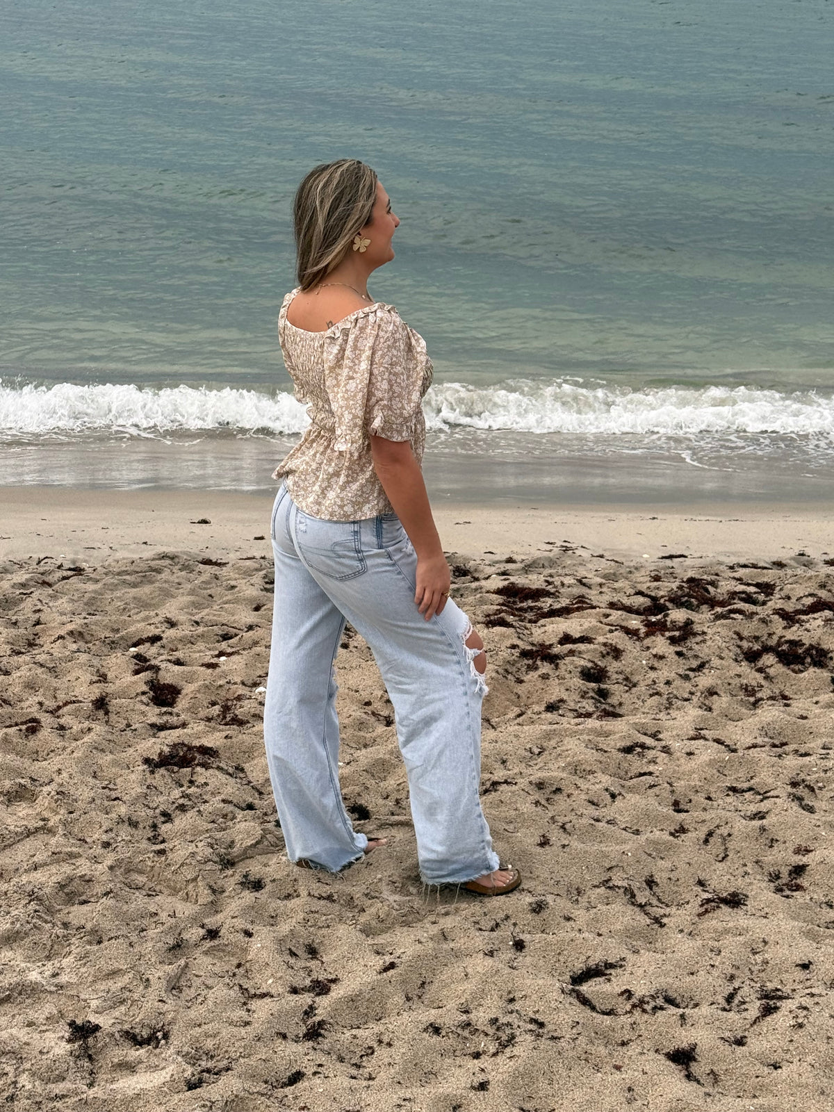 Back view of taupe and ivory floral blouse with smocked bodice and short flutter sleeves, styled with light wash jeans on the beach, Ardor Boutique.