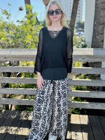 Woman wearing a black top and patterned pants standing on a wooden deck with greenery in the background