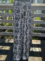 Black-and-white wide-leg pants with a bold floral print shown from the front, paired with white sneakers on a wooden deck.