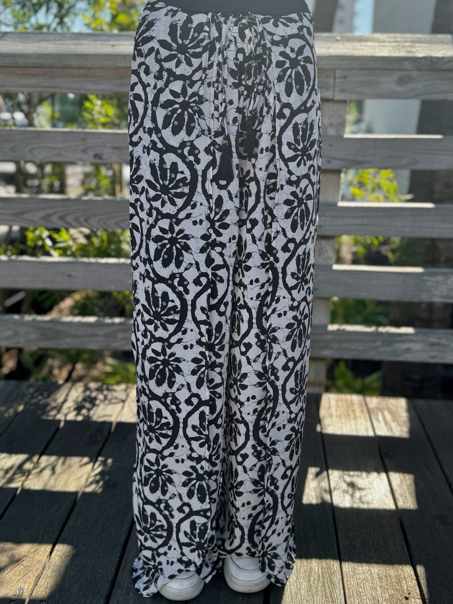 Black-and-white wide-leg pants with a bold floral print shown from the front, paired with white sneakers on a wooden deck.