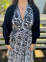 Woman wearing a black knit shawl layered over a black-and-white printed dress, standing outdoors on a wooden deck.