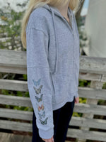 Person wearing a gray ardor hoodie with butterfly designs on the sleeves, standing outdoors.