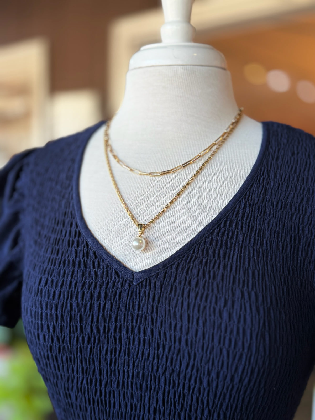 Gold rectangular link chain necklace layered with a gold pearl pendant necklace on a navy V-neck top at Ardor Boutique.