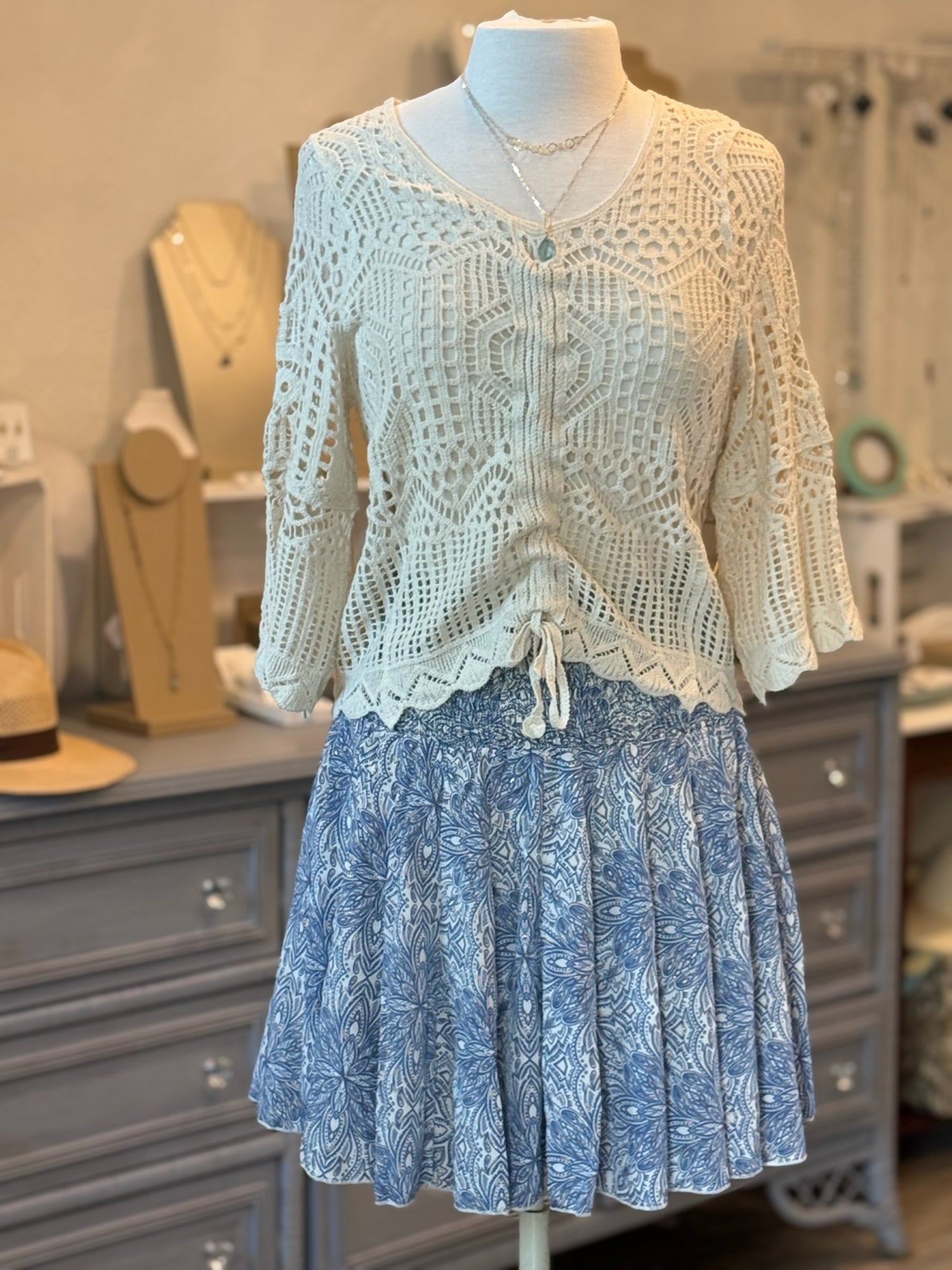 A beige crochet knit sweater with a v-neckline and adjustable ruched front displayed on a mannequin.