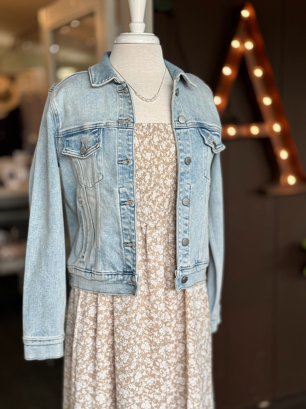 Light wash denim jacket layered over a beige and white floral dress with a delicate gold and pearl necklace, styled on a mannequin at Ardor Boutique.