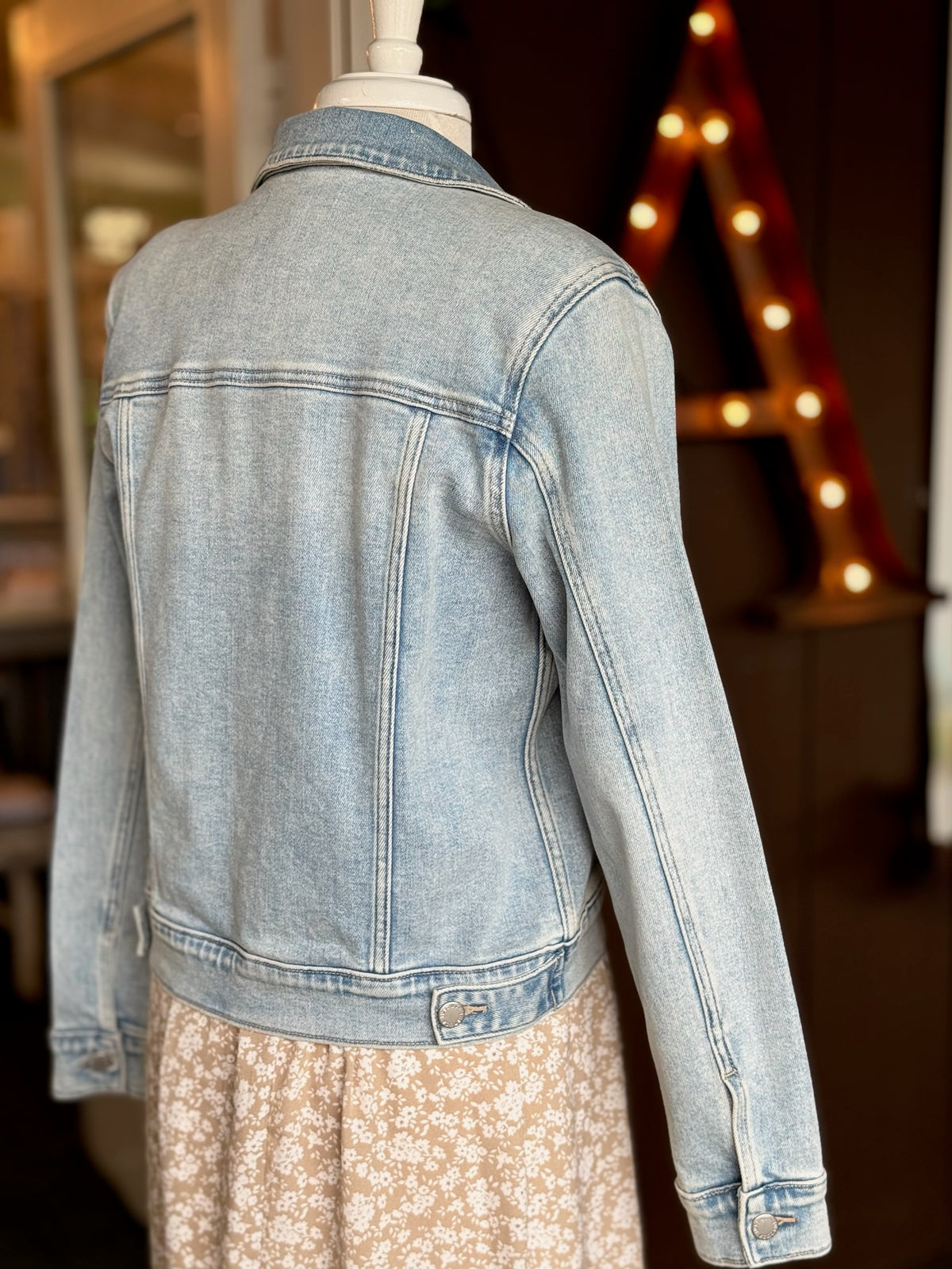 Back of the Light wash denim jacket layered over a beige and white floral dress styled on a mannequin at Ardor Boutique.