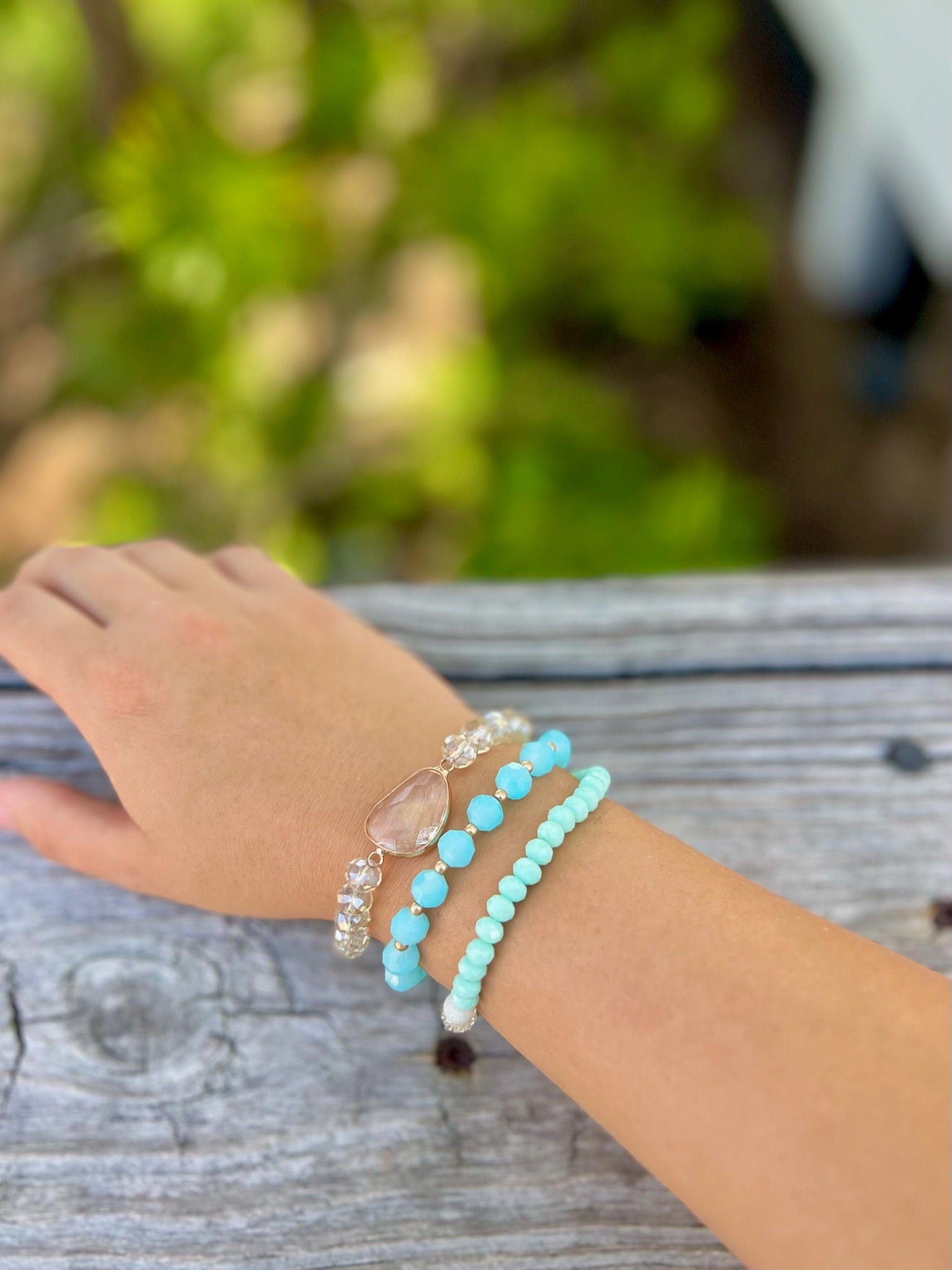 Stack of three beaded stretch bracelets in aqua, seafoam, and crystal tones worn on wrist outdoors on a wooden surface.