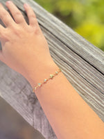 Hand wearing a gold bracelet with small stones, resting on a wooden surface with a blurred natural background.