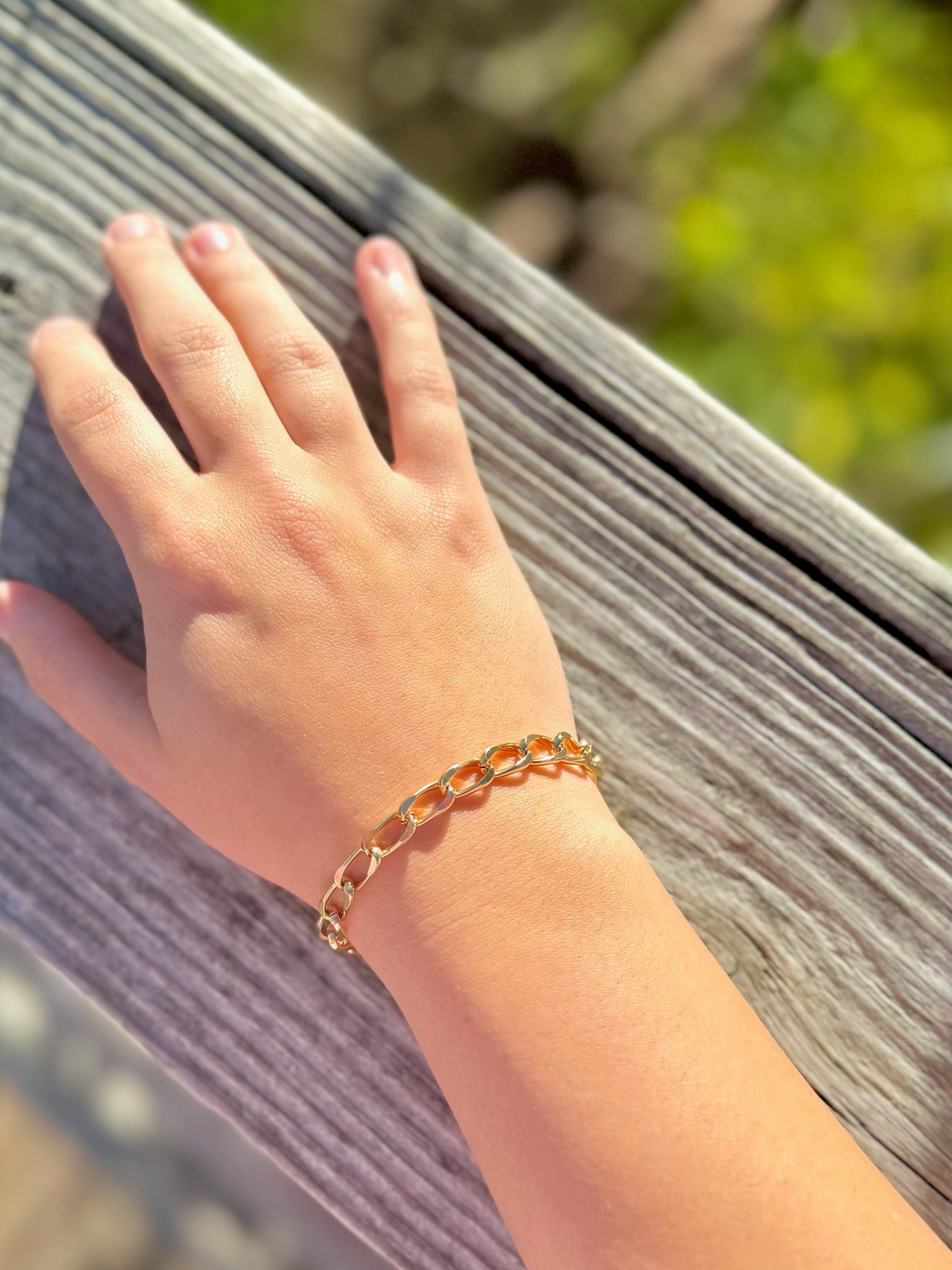 Hand wearing a gold chain bracelet against a blurred natural background