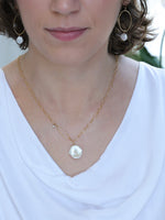 Woman wearing a handcrafted gold necklace with a large white Keshi freshwater pearl pendant and hammered oval accent, paired with coordinating gold hoop earrings featuring white pearl and moonstone drops. Styled with a white draped neckline top from the Nina Halls collection.
