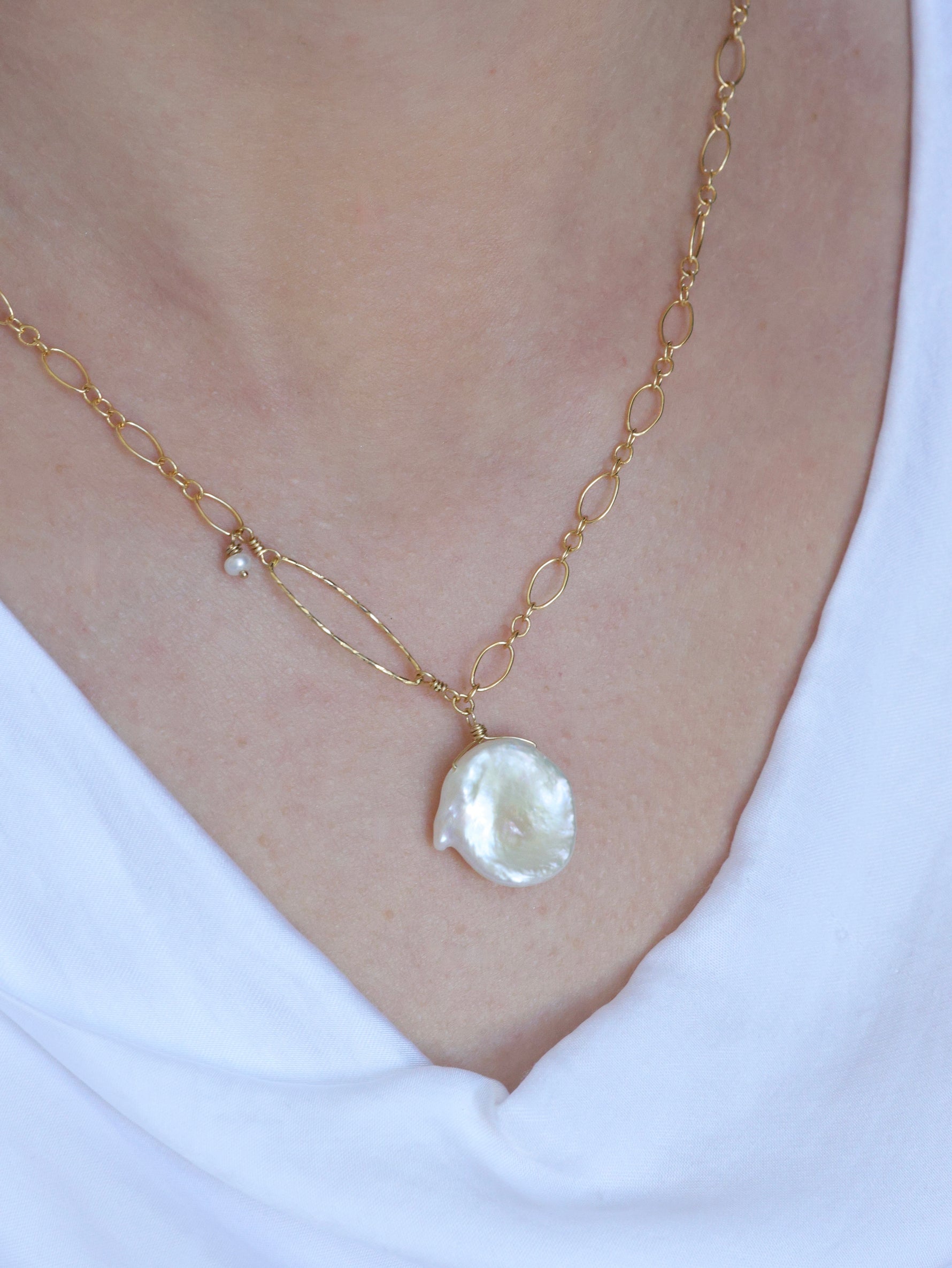 Close-up of a woman wearing a handcrafted gold necklace with a large white Keshi freshwater pearl pendant, featuring a hammered oval accent and small pearl charm. Asymmetrical chain design styled with a white draped neckline top from the Nina Halls collection.