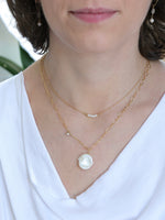 Woman wearing layered gold necklaces from the Nina Halls collection: a delicate pearl bar necklace and a larger white Keshi freshwater pearl pendant on a gold link chain with a hammered accent. Paired with drop earrings featuring pearl and moonstone. Styled with a white draped neckline top.