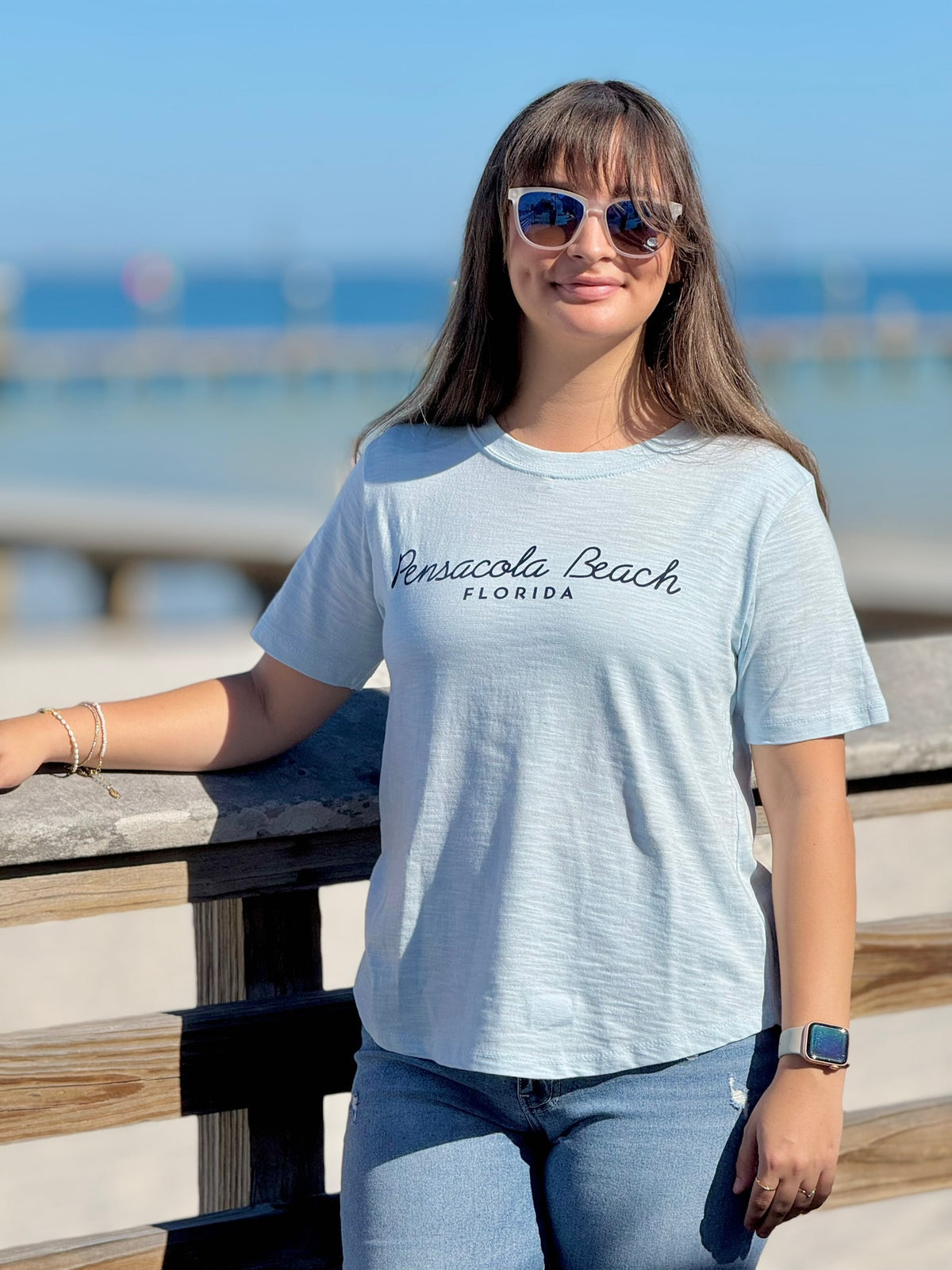 Woman wearing a light blue 'Pensacola Beach, Florida' t-shirt by the beach.