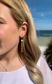 Mina gold earrings with a pearl at the top and a dangle gold accent hanging below the pearl in ear on model