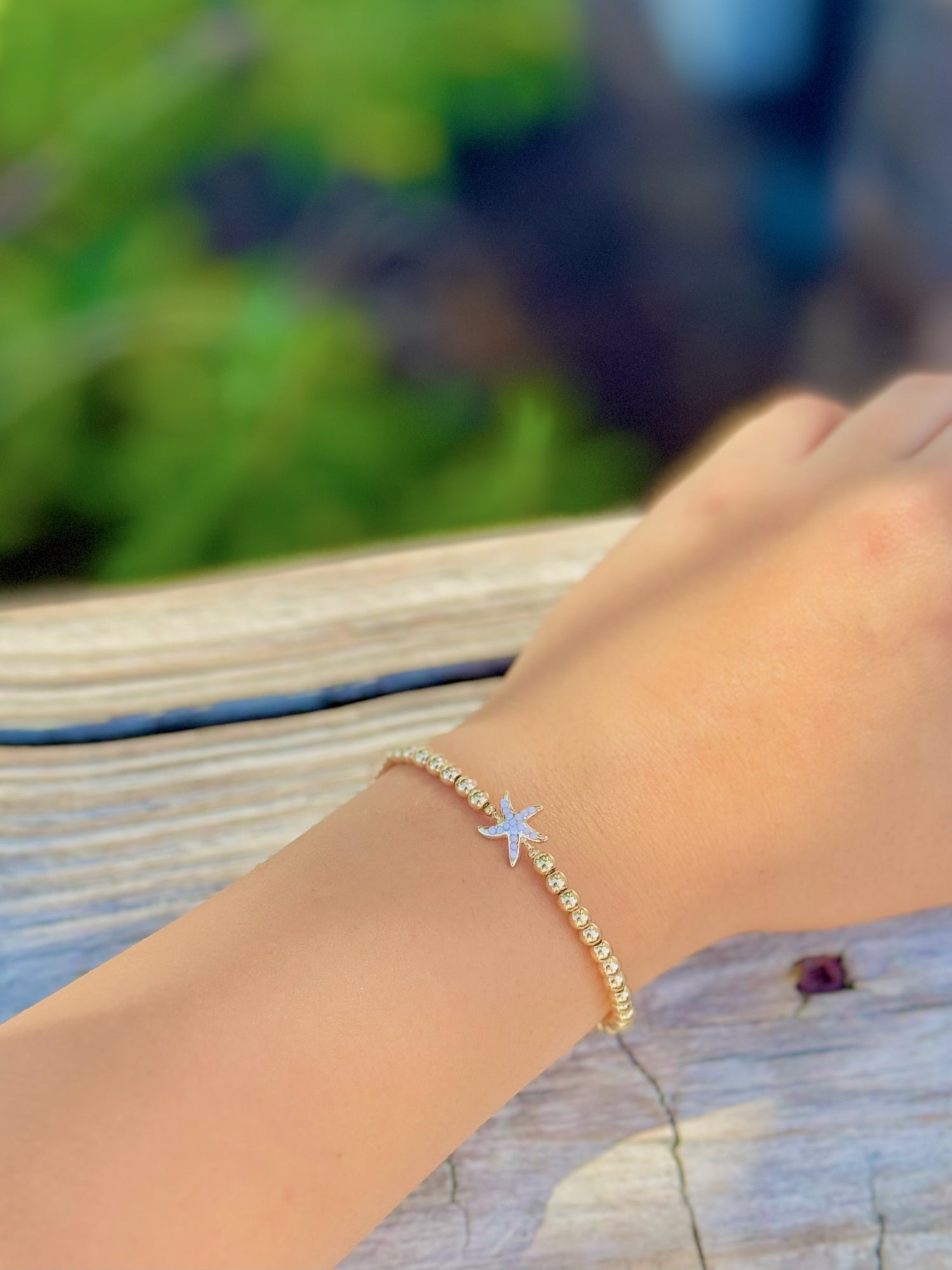 Hand wearing a gold bracelet with a starfish charm against a blurred natural background
