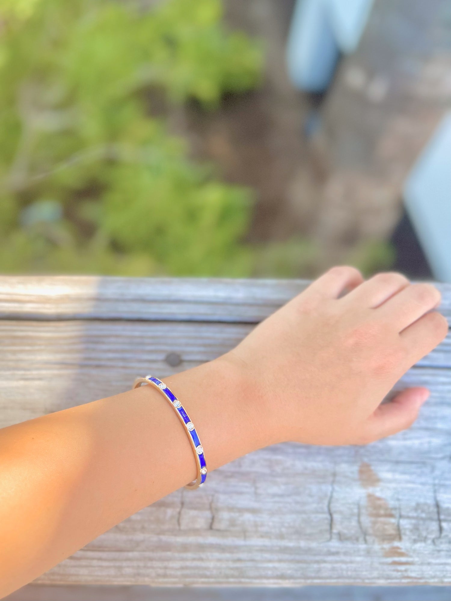 Hand wearing a blue and silver bracelet on a wooden surface with a blurred natural background